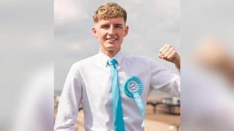 A young man with curly blond hair which is short at the sides is wearing a long-sleeved white shirt and a turquoise blue tie with a turquoise Reform UK badge. He is smiling and holding one fist up in the air.