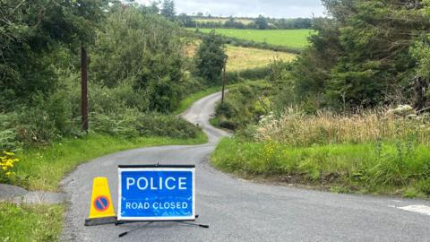A police sign place on the road says 'police road closed' and there is a yellow coin beside it. The road winds down through fields and shrubbery.  
