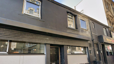 A dark-painted two-storey building on a narrow street, with a takeaway entrance at ground level and windows on the upper floor.