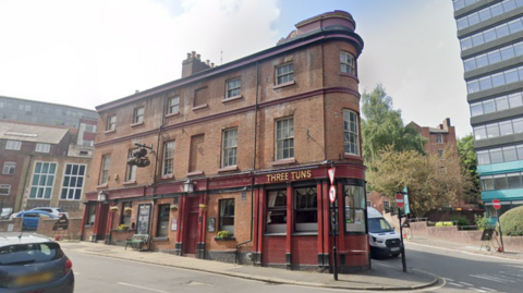 Street view picture of the pub. It is a distinctive 'flatiron' shaped pub with red painted detailing and the name font in gold.