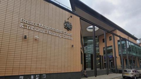 The exterior of the court, which is a large, modern building with a glass entryway and orange/brown brick-like cladding. The sign says 'Bristol Magistrates' Court & Tribunal Hearing Centre' on it.