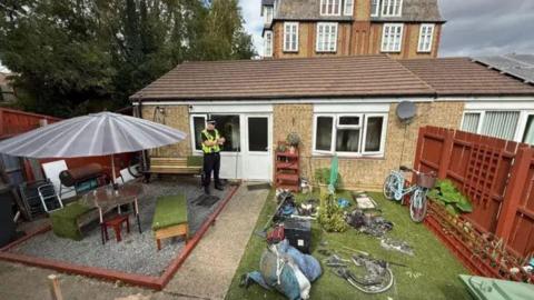 Back garden of a bungalow with green grass and garden furniture, with a police man dressed in his uniform, standing by the back door.