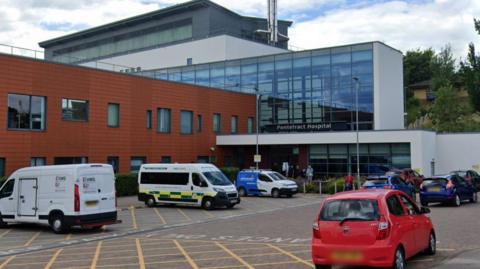 The outside of Pontefract Hospital which is a modern-looking building with brown tiles on one block and glass on another. There are three ambulances parked outside and a few cars.