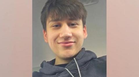 A portrait selfie of a young man wearing a black hoodie. He has brown hair with a fringe over his forehead and brown eyes. 