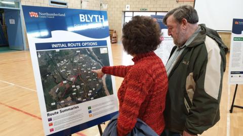 A man and a woman look at a panel showing options for the Blyth relief road on a map. The woman is pictured from behind and is pointing at something on the map. The man is side-on, next to her and is looking at the map. They are in a sports hall.