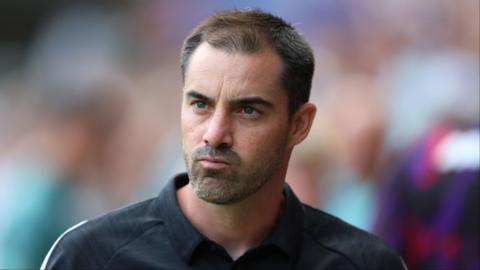 Ruben Selles pouts as he looks out to the distance. He wears a black polo shirt and stubble.