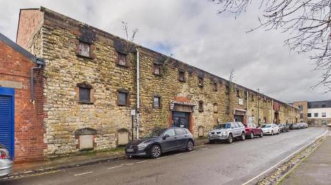 The outside of an old music venue. It is a stone brick, warehouse-style building. It looks quite run down, with boarded up windows. There are cars parked alongside it.