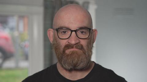 Keith Macaldowie, wearing a round neck black t-shirt, stares straight at the camera with a serious expression on his face. He is bald, wearing dark-rimmed glasses and has a bushy ginger and grey beard and moustache. The image was captured in his home with white walls and a car bumper - seen through a window - visible in the background.