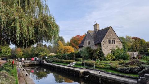 A house next to a canal under a mostly cloudy sky. Trees can be seen in the background with orange and yellow autumnal colours