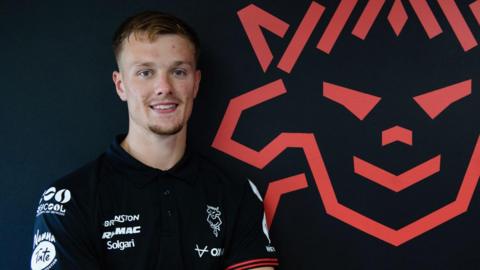 Oscar Thorn wearing a Lincoln City top, he is smiling in front of the Lincoln City badge which is red on a black wall. 
