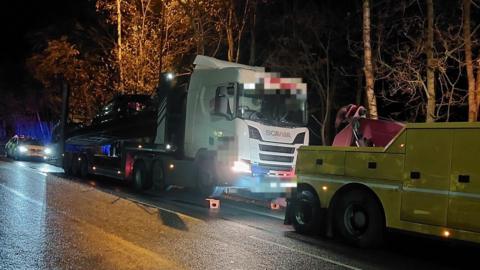 A white cabbed car transporter lorry being towed by a yellow rescue truck. Behind it is a police car. It is night time.