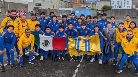 A team photo of players and coaches all wearing their club’s tracksuit tops and holding flags