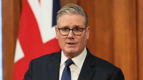 Headshot of Starmer who speaks and wears black framed glasses. He stands in front of a wooden panelled wall and a British flag that is curled up