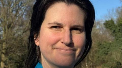Donna Edmunds, smiles at the camera. She has dark hair and is stood with a woodland behind her 