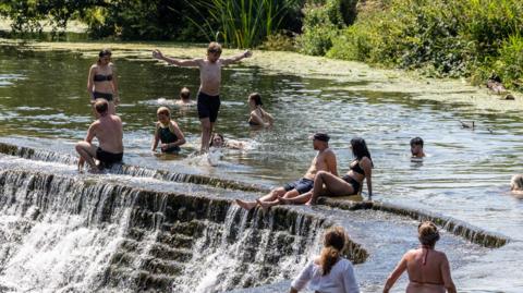 A group of wild swimmers in the River Avon at Warleigh Weir. Some people are swimming while others are standing in the water or sitting by the edge of the weir. One man has his arms outstretched as he stands in the water.