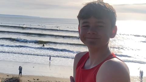 Oscar is smiling at the camera as he stands with Saunton Sands stretching out behind him on a sunny day. The beach and sea are busy with surfers, swimmers and walkers.