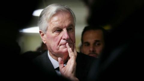 A headshot of Michel Barnier holding his hand to his mouth looking pensive