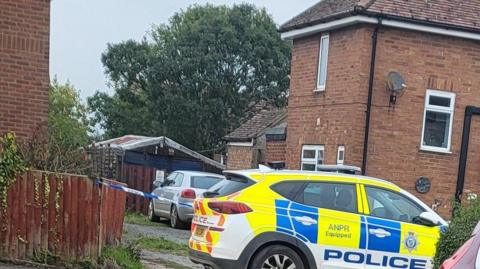 Close up shot of a police vehicle at the scene - it has yellow and blue markings and the words "ANPR equipped" and the emblem of Lincolnshire Police on the side. A house and another vehicle can be seen in the background. 