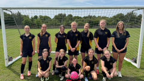 A photo of 12 members of a female football team, the front row sitting and the back row standing, in front of a goal. They are wearing black and yellow football kids. The girl in the middle of the front row is holding a pink football. They are all looking at the camera and smiling.