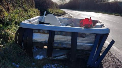 Abandoned hot tub at side of a road - it is in a poor state and filled with other rubbish.