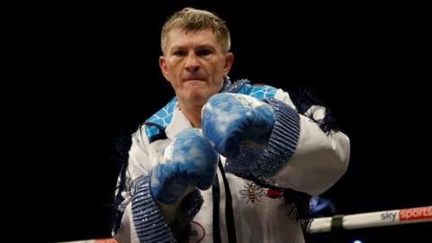 Ricky Hatton stands in a boxing ring. He is wearing a white tasselled suit and blue boxing gloves and punching the air. 