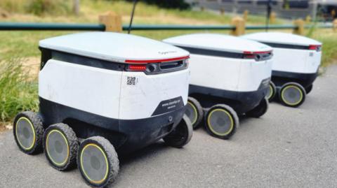 Three white robots with black wheels parked up alongside each other on tarmac.