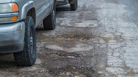 A grey car is stationary next to around four or five large potholes.