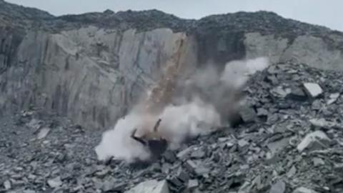 Dust and debris falling from a grey slate cliff, with a bank of slate ruble leading up to the cliff
