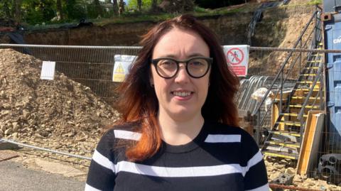 A woman in a stripey top and black glasses with red shoulder-length hair standing in front of an excavated bank which is fenced off. 