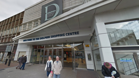 A screenshot from Google street view showing the front entrance to a shopping centre. It has a row of glass doors and a large 'D' as a logo.