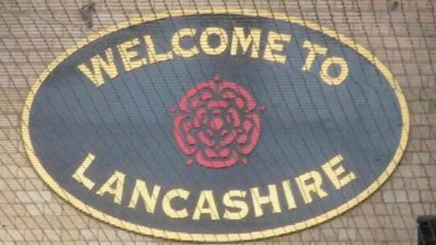 Oval sign saying "welcome to Lancashire" with a red rose in the middle. the writing is gold on a black background