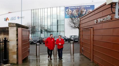 Two railway workers in red jackets walk through the new station entrance. A brown-panelled wall on the right has letters mounted to it that read Newcastle, next to a British Rail logo. Behind them is the large glass and silver metal International Centre for Life.