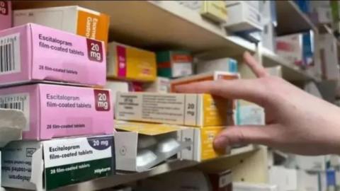 Boxes of medication on a shelf in a chemist, with a hand reaching for two boxes.