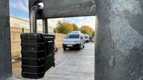 A padlock, closed, on to a gate latch at Parravani's, with chiller vans parked in a yard visible beyond.