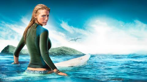 Blake Lively sits on a surfboard in the middle of large body of water. There's an island in the background