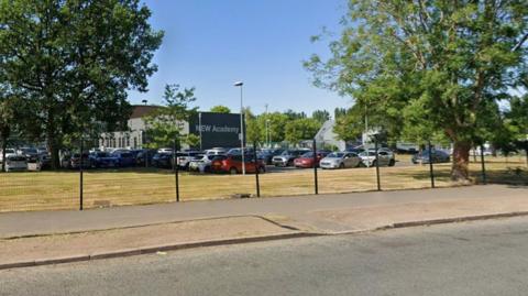 Dozens of cars parked in front of a large building situated behind gates on a sunny day.