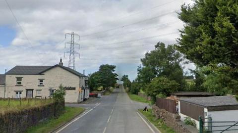 A country road showing a junction between a pub on the corner and on the opposite side a tree-lined road