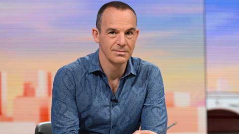 Martin Lewis, wearing a blue shirt, sitting at a desk