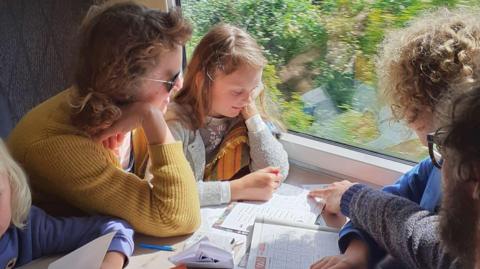 A family sitting by a table on a train. They are looking at workbooks on the table.