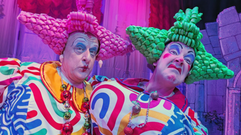 Two panto dames are wearing brightly-coloured costumes and exaggerated make-up. The dame on the left is wearing a bright pink pointy plastic wig. The dame on the right is wearing green pointy plastic hair.