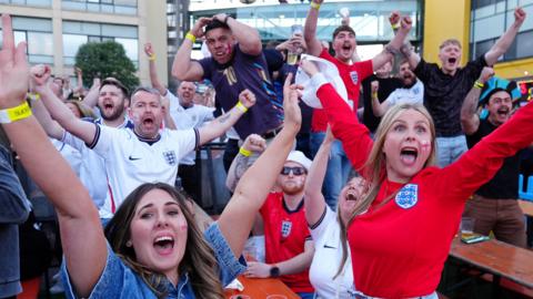 A crowd of England supporters - men and women - dressed in red and white kit shirts, some wearing St George's Cross face paint, and cheering with their fists in the air.