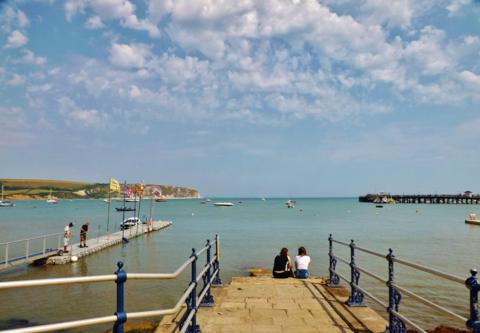 Two people sitting on the end of a short slipway with a pontoon, harbour and pier in the background