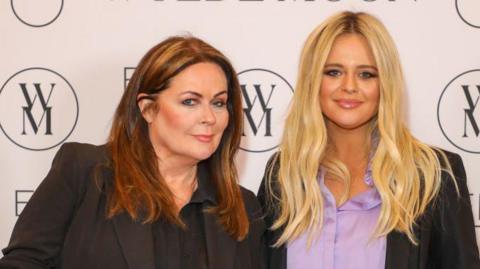 Kate Robbins, with brown hair, is stood next to Emily Atack, with blonde hair, at a press event. There is a white wall behind them with a brand logo that looks like a W and M joined together in a circle.