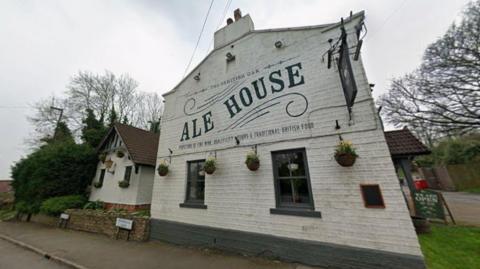 Front exterior of The British Oak pub, a white-painted brick building with large “Ale House” lettering on the façade, hanging baskets and a roadside setting bordered by trees.