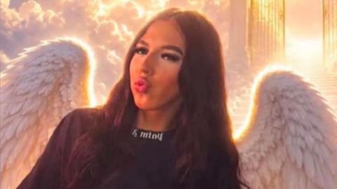 Victoria Niebylska, who has long, wavy brown hair with a centre parting and is pouting. She is wearing a black tshirt with white writing on the collar and angel wings have been added to the picture, which are glowing against the sky