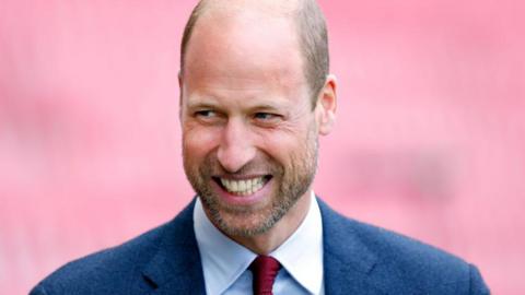Prince William, wearing a navy suit and red tie, smiles