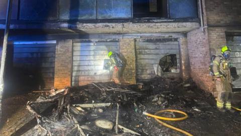 A block of flats severely damaged by fire. Firefighters an be seen assessing the damage. The fire is out.