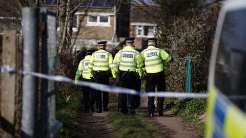 A group of uniformed Sussex Police officers walk down a lane with their backs to us. In the foreground the edge a police van can be seen on the right hand side. The land is cordoned off with police tape.