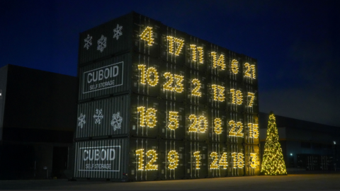 A night-time image of a structure made from shipping containers. There are six columns containing four containers each. On the front of the containers are numbers, lit up. On the right of the containers is a large christmas tree