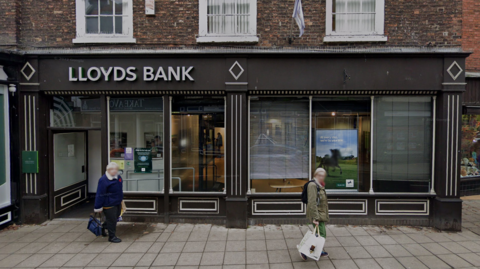 The exterior of Lloyds Bank in Horncastle. The high street building is painted black and there are two people, with their faces blurred, walking in front of the building.